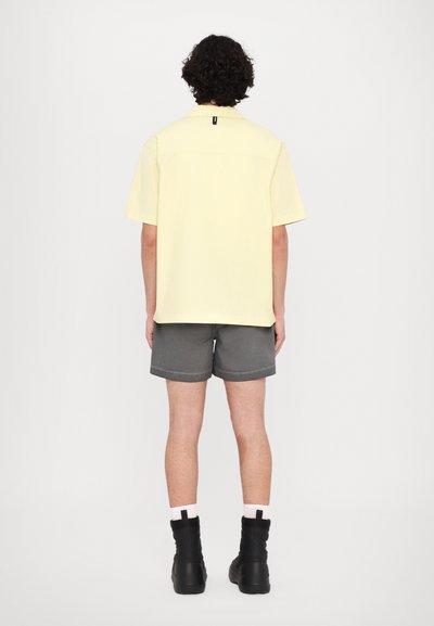 Camisa amarilla de manga corta con corte recto, combinada con pantalones cortos grises. Botas negras con suela gruesa completan el atuendo.