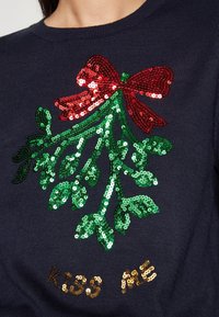 Marineblauwe trui met een met pailletten geborduurde mistletoe in rood en groen, met "KISS ME" in gouden pailletten geschreven.