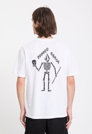 Personne portant un t-shirt blanc avec un motif squelette noir tenant un crâne et une lance, texte "Volcom Stone" au-dessus de la figure.