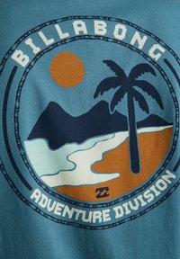 Modrá látka s kruhovým logom zobrazujúcim palmovník, hory, slnko, pláž a text „Billabong Adventure Division“.