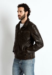 Man met donker krullend haar en baard, gekleed in een donkerbruine leren jas en lichtblauwe spijkerbroek, staand met handen in de zakken.