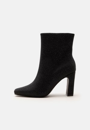 Bottine noire recouverte de petites décorations texturées avec un bout pointu et un talon haut et épais, sur un fond uni.