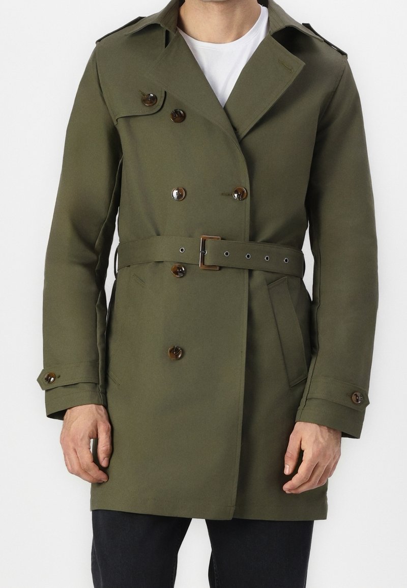 Homme portant un trench-coat croisé vert olive avec ceinture, poignets boutonnés, et un t-shirt blanc à col rond en dessous.