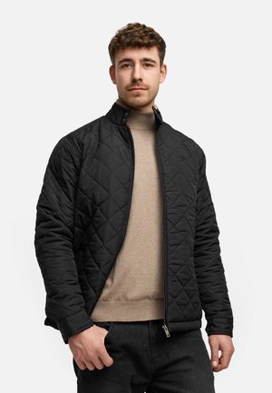 Svart quiltet jakke med ståkrage, glidelås og subtil logo-detalj, båret over en lys beige turtleneck genser.