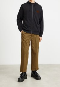 Felpa nera con zip, cappuccio regolabile e un piccolo patch logo marrone, abbinata a pantaloni dritti marroni e scarpe platform nere.