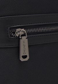 Närbild av en svart texturerad väska med ett metallblixtlås med varumärket "Valentino" ingraverat.