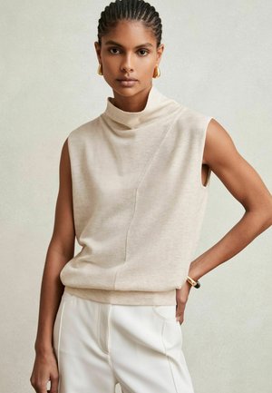 Beige sleeveless top with a high neckline, made of soft fabric, featuring a diagonal seam detail. Worn with white pants and minimal accessories.