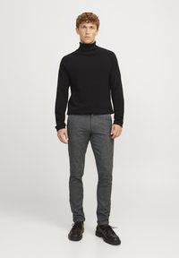 Schwarzer gerippter Rollkragenpullover, kombiniert mit strukturierten grauen Hosen und schwarzen Schnürschuhen. Model steht vor einem weißen Hintergrund.