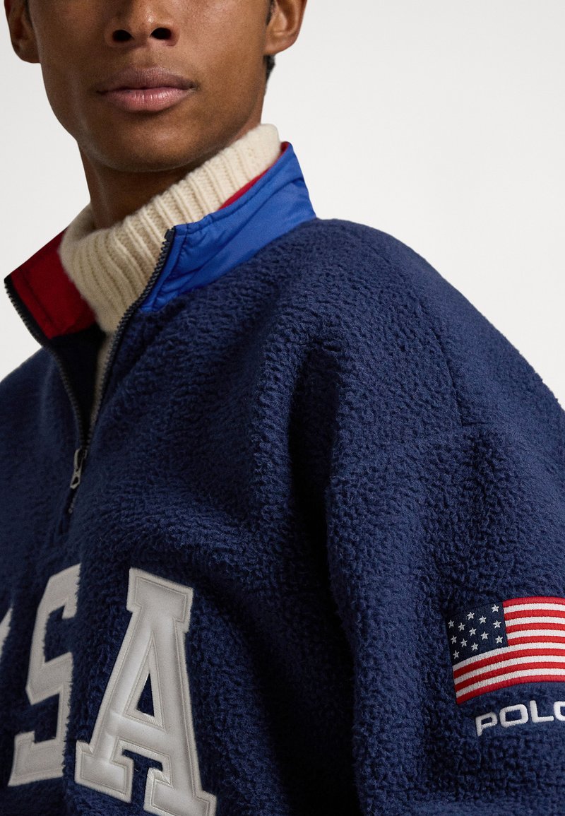 Pull-over en polaire bleu marine avec une fermeture éclair au quart, doté d'un col de pull crème, d'un lettrage blanc "USA" et d'un petit patch drapeau américain sur la manche.