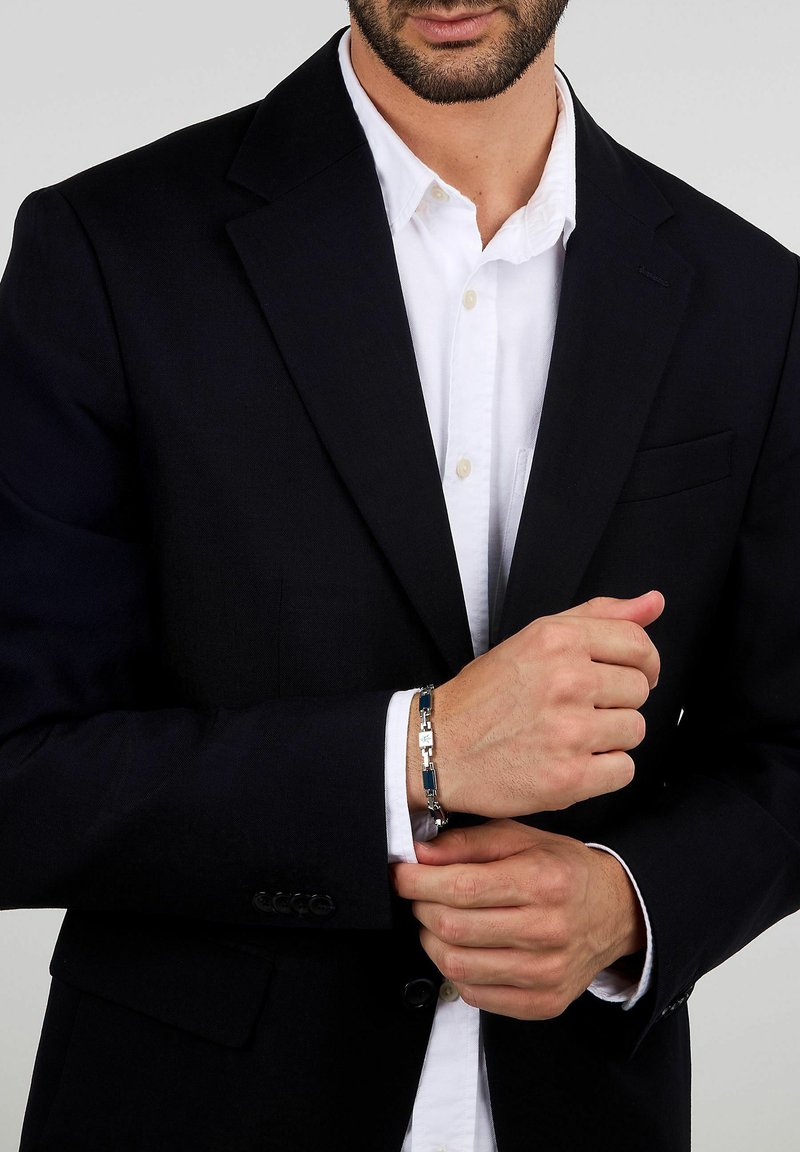 Silverfärgad metallarmband med blå accenter, med en geometrisk design. Bärs på handleden medan man är klädd i en svart kostym och vit skjorta.