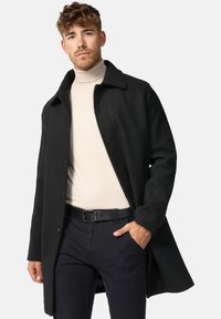 Sort uldfrakke med spids krave, lange ærmer og knapper foran. Båret over en lys cremefarvet turtleneck, kombineret med mørke bukser og et bælte.