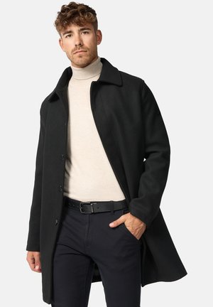 Sort uldfrakke med spids krave, lange ærmer og knapper foran. Båret over en lys cremefarvet turtleneck, kombineret med mørke bukser og et bælte.