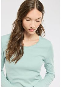 Haut à manches longues vert clair avec une texture côtelée et un col rond. La coupe est ajustée, mettant en valeur la silhouette.