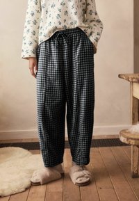 Fekete-fehér kockás, laza nadrág, állítható derékpánttal, bézs bolyhos papuccsal kombinálva, egy fa padlón viselve.