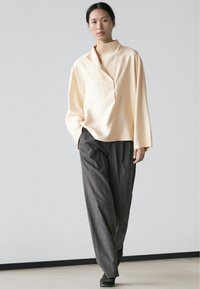 Vrouw loopt in loszittende crèmekleurige blouse met hoge kraag en grijze broek met wijde pijpen tegen een effen achtergrond.