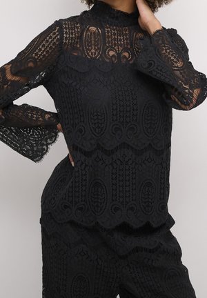 Femme portant un chemisier en dentelle noir à col montant et un pantalon assorti avec des motifs floraux et géométriques complexes.