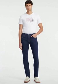 Pantalones vaqueros azul marino de corte ajustado, combinados con una camiseta blanca que presenta un gráfico rosa de "GUESS JEANS". Zapatillas en gris claro y beige.