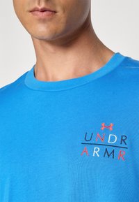 Mėlynas medvilninis marškinėliai su apvaliu kaklu; priekyje siuvinėjamas "UNDER ARMOUR" logotipas raudona, tamsiai mėlyna ir balta spalvomis.