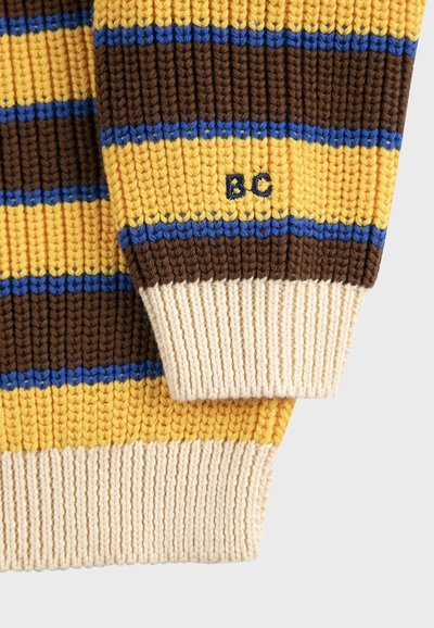 Strikket sweaterærme med horisontale striber i gul, brun, blå og beige, broderet med bogstaverne "BC" på den gule stribe.