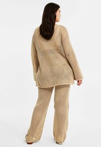 Beige meshstickad tunikakjol och byxor, med lös passform, långa ärmar och strukturerad mönstring. Designad för komfort och andningsförmåga.