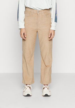 Person iført beige kordfløjlsbukser, hvid langærmet skjorte med grafiske ærmer og hvide sneakers, der står mod en ensfarvet baggrund.