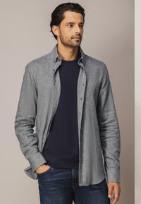Grijze button-up shirt met een gestructureerde stof, lange mouwen en een relaxed fit, gedragen over een marineblauw t-shirt, gecombineerd met donkere jeans.