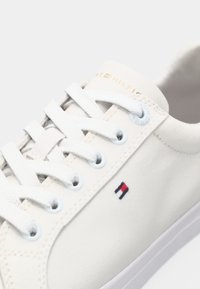 Gros plan sur une sneaker blanche avec des lacets blancs, un petit logo rouge, blanc et bleu sur le côté, et un branding subtil sur la languette.
