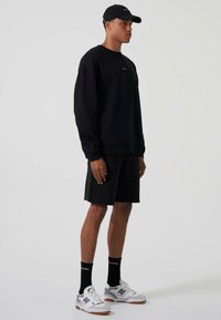 Felpa nera con polsini a costine, abbinata a pantaloni corti neri. Gli accessori includono un cappellino nero e sneakers bianche con accenti grigi e marroni.