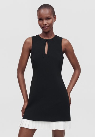 Femme mince portant une robe noire sans manches avec une petite découpe devant et un ourlet plissé blanc, debout devant un fond clair uni.
