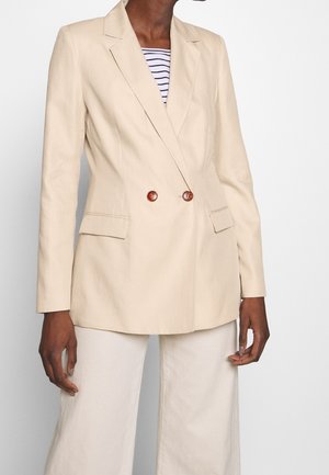 Blazer beige à double boutonnage avec deux boutons marron sur une chemise rayée, porté avec un pantalon large beige clair.