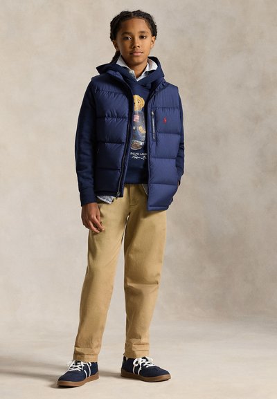 Ragazzo che indossa un gilet imbottito blu navy, una felpa con cappuccio blu navy con grafica orso, pantaloni beige e sneaker blu navy, in piedi davanti a uno sfondo semplice.