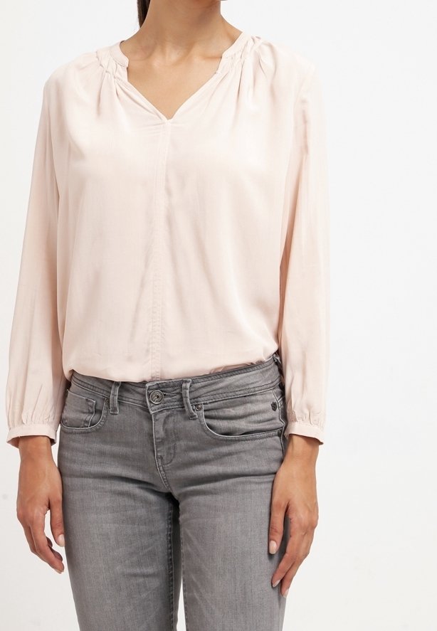 Blouse rose pâle à manches longues avec un col en V et des détails froncés au niveau de l'encolure, associée à un jean gris. Tissu lisse avec un léger éclat.