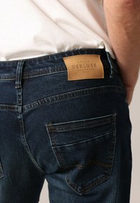Jean en denim bleu foncé avec une étiquette en cuir « Deeluxe » à l'arrière de la taille et des poches arrière classiques avec des détails de couture subtils.
