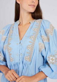  Lichtblauwe blouse met opgeblazen mouwen, versierd met ingewikkelde witte en gouden borduursels, een knopenlijst en een gerimpelde taille. 