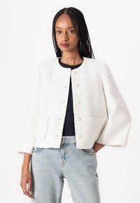 Weiß strukturierte Jacke mit rundem Ausschnitt, Knopfverschluss, zwei Fronttaschen und weiten Ärmeln, kombiniert mit blauen Jeans.