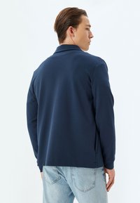 Camisa polo de manga larga en color navy con corte recto, textura suave y aberturas laterales. Lleva puesta unos vaqueros de color azul claro, vista desde la espalda.