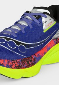 Κοντινό πλάνο ενός μπλε αθλητικού παπουτσιού με μοβ κορδόνια, μαύρο και λευκό λογότυπο, νεον κίτρινη σόλα και κόκκινα υφασμένα διακοσμητικά στη μεσαία σόλα.