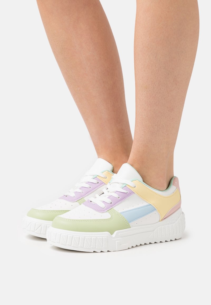 Personne portant des baskets blanches avec des panneaux pastel verts, violets, jaunes, bleus et roses, debout sur un fond blanc.