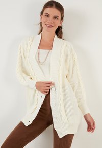 Cárdigan blanco de punto con detalles de trenzado, cierre de botones y puños acanalados. Llevado sobre una blusa clara y pantalones marrones.