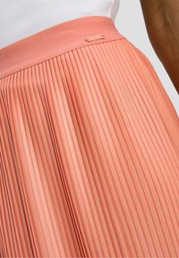 Falda plisada en tejido coral suave con una cinturilla ancha y una pequeña etiqueta con el logo metálico. Presenta pliegues verticales y una textura lisa.