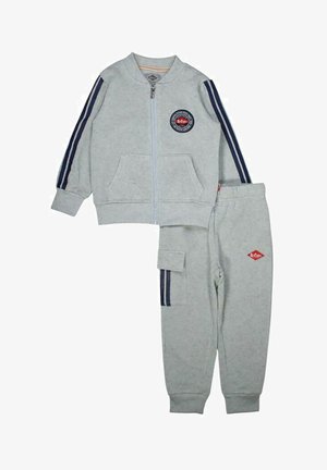 Ensemble de survêtement gris clair en tissu doux. Veste zippée avec des rayures bleu marine et un logo rond ; pantalon assorti avec une poche latérale et des rayures.