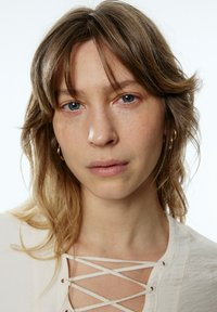 Nahaufnahme einer Person mit hellbraunen, lockigen Haaren und blauen Augen. Sie trägt ein beiges Oberteil mit einem überkreuzten Spitzenmuster.