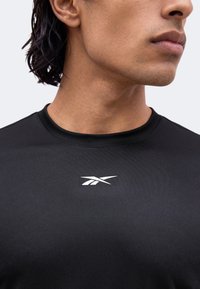 Jeune personne au teint moyen porte un t-shirt noir avec un logo sportif blanc sur la poitrine, tête tournée sur le côté.