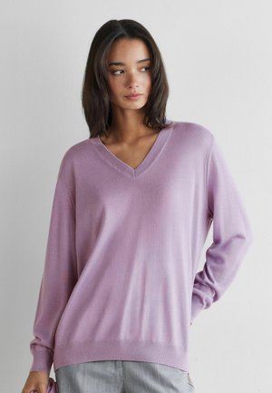 Ung kvinde med mørkt hår iført en løs lilla V-hals sweater og grå bukser, stående mod en ensartet hvid væg.