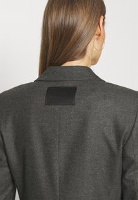 Szary żakiet z teksturowanej tkaniny, z wyciętym kołnierzem i czarną skórzaną łatą z napisem "HUGO" na górnej części pleców.