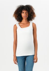 Mujer con cabello rizado que lleva una blusa sin mangas blanca y jeans azules, de pie contra un fondo blanco liso.