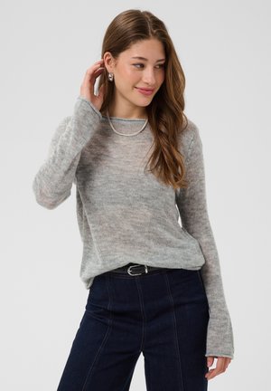 Femme aux longs cheveux bruns portant un pull gris clair, un jean foncé, un collier en argent et des boucles d’oreilles, souriante et touchant ses cheveux.