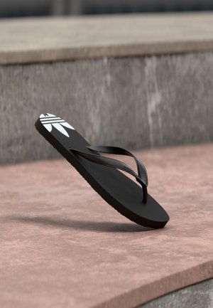 Zwarte teenslipper met wit logo op de zool, zwevend boven een roze stenen oppervlak en een grijze stenen trap als achtergrond.