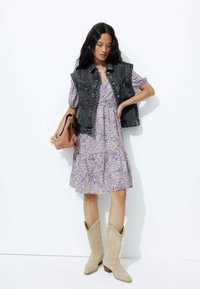 Veste en jean sur une robe à volants à motif lilas ; associée à des bottes de cow-boy beiges et portant un sac beige. Look simple et contemporain.