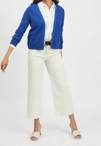 Modrý pletený svetr na knoflíky s volným střihem, bílá košile s límečkem, krémové široké kalhoty s vysokým pasem a béžové sandály s otevřenou špičkou.